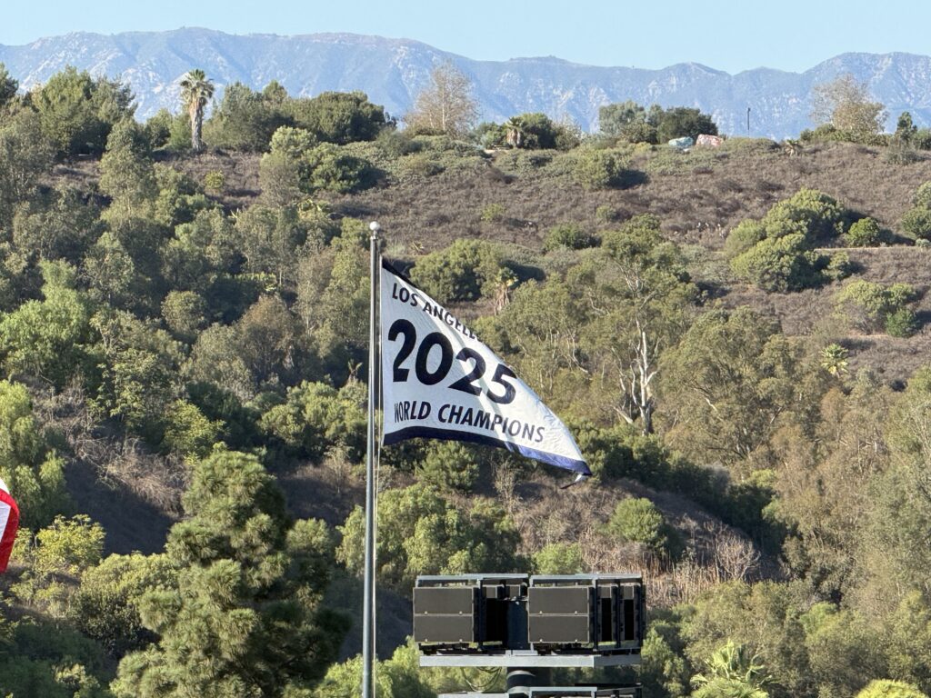 A white "Los Angeles 2025 World Champions" flag flies above the hills beyond Dodger Stadium, with the San Gabriel Mountains in the background.