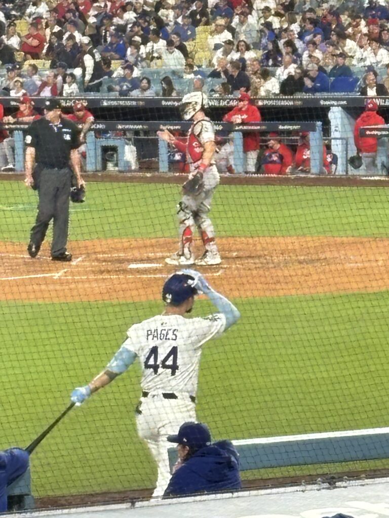 Dodgers outfielder Andy Pages, No. 44, adjusts his helmet near the on-deck area as a Cincinnati Reds catcher and the umpire stand at home plate.