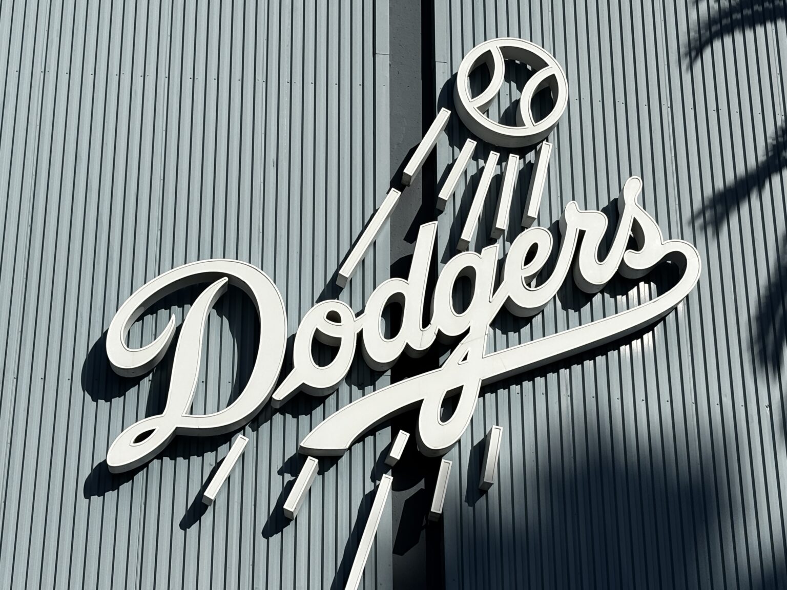 Close-up of the white Dodgers script logo and flying baseball mounted on the corrugated exterior wall of Dodger Stadium.