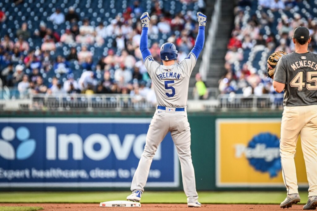 Freddie Freeman in a gray Dodgers road uniform with number 5 standing on a base with both arms raised.
