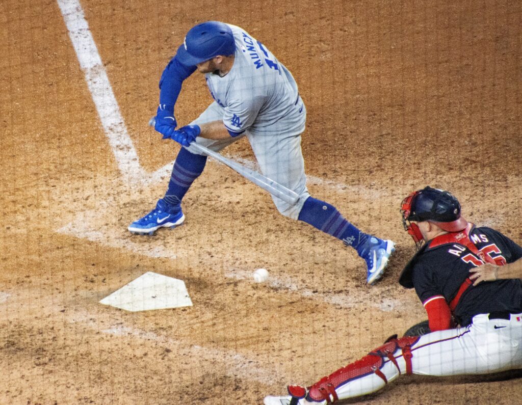 Max Muncy in a gray Dodgers road uniform reaching for a low pitch as the Nationals catcher dives beside him.