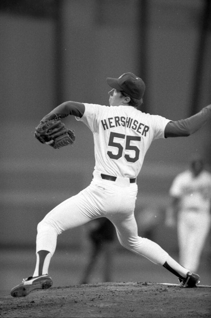 Black-and-white photo of Orel Hershiser, number 55, striding forward mid-pitch, seen from behind.