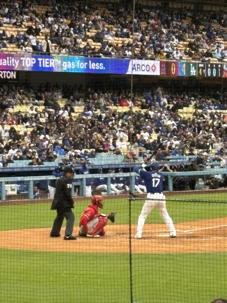Shohei Ohtani in a blue Dodgers No. 17 jersey waits for a pitch as a Los Angeles Angels catcher and the umpire set up behind the plate.