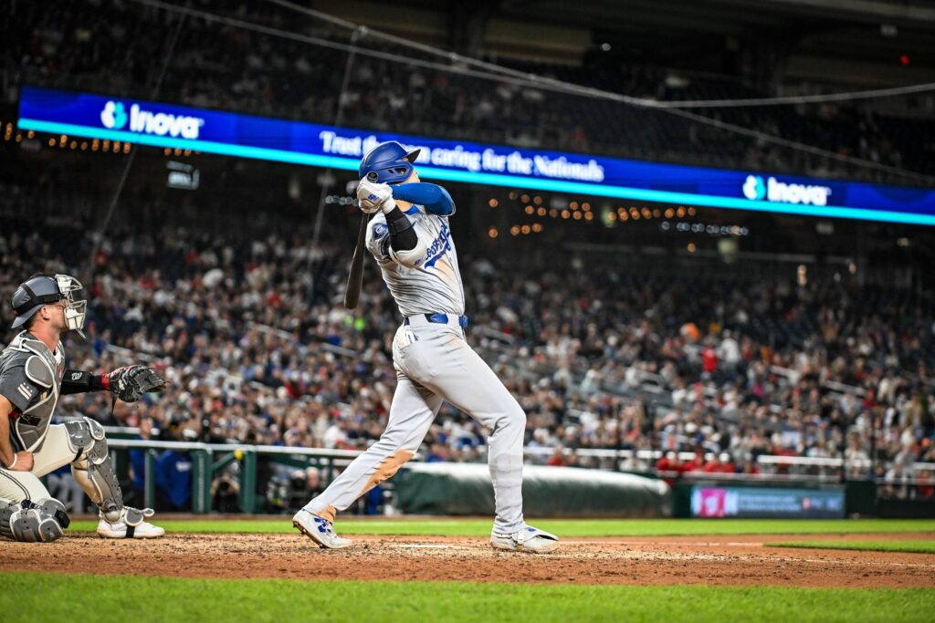 Shohei Ohtani in a gray Dodgers road uniform swinging at home plate in front of a packed crowd.