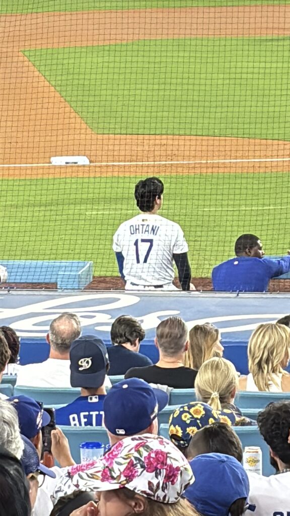 Shohei Ohtani, in a white No. 17 jersey, sits at the edge of the Dodgers dugout facing the field, with fans in the foreground.